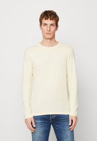 Kremfarget langermet T-skjorte i mykt stoff med rund hals, båret med blå jeans. Enkel design, uten synlige mønstre eller aksenter.