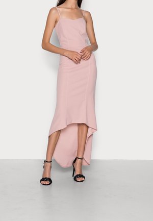 Vrouw draagt een lichtroze mouwloze high-low jurk met dunne bandjes en zwarte gevlochten hoge hakken sandalen, staand tegen een effen muur.