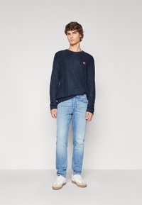 Pull en tricot marine avec un motif texturé, encolure ronde et petit logo; associé à un jean bleu clair et des baskets blanches.