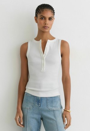 Mujer con pelo trenzado, vestida con una blusa blanca sin mangas abotonada y vaqueros azul claro, de pie frente a una pared blanca lisa.