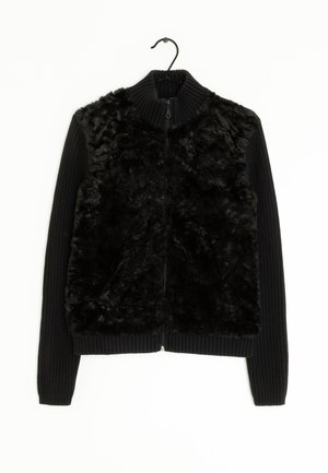 Veste noire zippée avec un devant en fausse fourrure et des manches en tricot côtelé. Le col est également côtelé pour une texture supplémentaire.