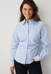Chemise bleu clair à boutons avec un col structuré, des fronces à la taille et des manches longues, portée avec un jean foncé.