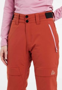 Red, waterproof ski pants with two front pockets, adjustable waistband, and a side zip pocket in pink. Logo displayed on the lower leg.