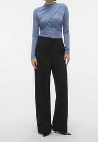 Pitkähihainen sininen paita, jossa on laskeutuva muotoilu, yhdistettynä korkeavyötäröisiin mustiin leveälahkeisiin housuihin ja mustiin, teräväkärkisiin korkokenkiin.