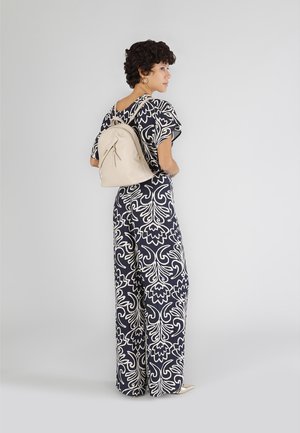 Beige leren rugzak met ritssluiting, gladde textuur en bovenhandgreep, gecombineerd met een marineblauwe en witte gestreepte jumpsuit.