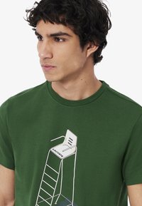 Vihreä puuvillapaita, jossa on valkoinen pelastustornin grafiikka ja 'LACOSTE' -teksti, lyhyet hihat ja klassinen pyöreä pääntie.
