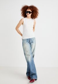 White sleeveless top paired with loose light blue jeans and red high-heeled shoes. Model wears large black sunglasses; medium curly hair.