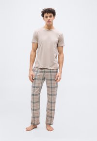Νέος άνδρας όρθιος, ξυπόλητος, φορώντας μπεζ μπλούζα και μπεζ πιτζάμες με καρό σχέδιο, μπροστά σε ανοιχτό γκρι φόντο.
