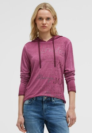 Young woman with blonde hair wearing a purple long-sleeve hoodie with printed text and blue jeans, standing against a plain background.