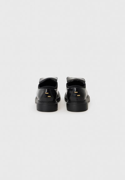 Chaussures en cuir noir avec une finition lisse, bout rond et semelle en caoutchouc noir épaisse. Présente des accents dorés minimalistes à l'arrière.