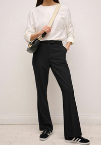 Pantalons évasés noirs au tissu lisse, associés à une chemise blanche à manches longues. Un petit sac bandoulière noir et des baskets noires complètent le look.