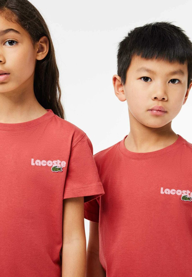 Rote Baumwoll-T-Shirts mit dem "Lacoste"-Logo in Pink und einem Krokodilsymbol, mit einem klassischen Rundhalsausschnitt und kurzen Ärmeln.