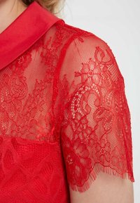 Vestido rojo con un cuerpo de tela sólida y mangas cortas de encaje translúcido, que presenta patrones florales y acentos de flecos a lo largo de los bordes de las mangas.