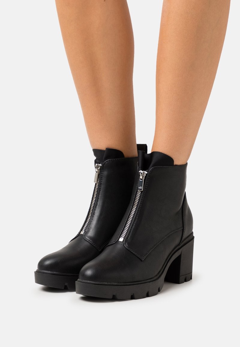 Bottes cheville en cuir noir avec fermetures éclair argentées à l'avant, talons épais et semelles crantées portées sur des jambes nues sur un fond blanc.