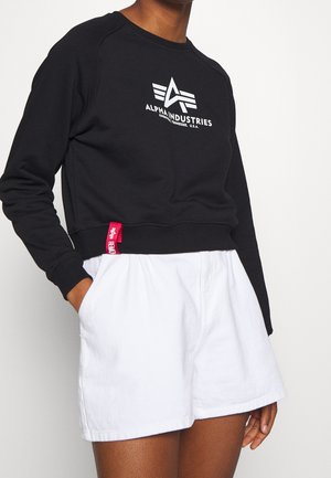 Person iført en sort Alpha Industries sweatshirt med logo og hvide højtaljede shorts, hænder i lommerne mod en ensfarvet baggrund.