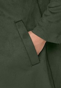 Abrigo verde oliva con una textura suave, que cuenta con un bolsillo lateral y un diseño elegante; las costuras visibles resaltan los bordes.