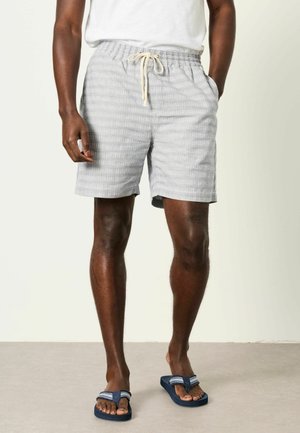 Mann trägt hellgraue gestreifte Shorts mit Kordelzug, ein weißes Hemd und blaue Flip-Flops und steht auf einem schlichten Boden.