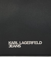 Juodas tekstūruotas odos paviršius su sidabriniu logotipo ženkleliu, ant kurio užrašyta "KARL LAGERFELD JEANS" ryškiais, moderniais šriftais.
