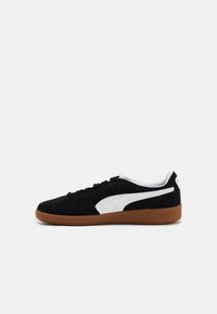 Sneaker in suede nera con accenti bianchi, suola piatta in gomma marrone, punta rotonda e chiusura con lacci. Texture liscia con elementi di design a contrasto.