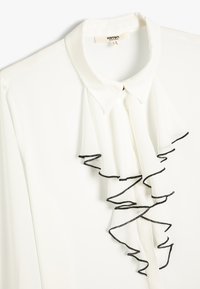 Blusa blanca con un collar estructurado, con un frente con volantes acentuado con un ribete negro. Fabricada en tela ligera, de textura suave.