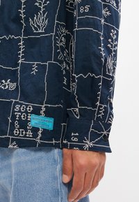 Ναυτικό πουκάμισο με λευκή γεωμετρική κέντηση, κουμπιά στους καρπούς και τιράντα μάρκας σε τυρκουάζ. Το ύφασμα έχει υφασμένη όψη.