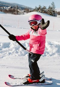 Kind dat op een besneeuwde helling skiet, gekleed in een roze geïsoleerde jas, zwarte broek, roze helm en skibril, en een skistok gebruikt ter ondersteuning.