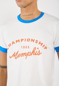 Bijela pamučna majica s plavim ovratnikom i rukavima, s narančastim natpisom: "CHAMPIONSHIP 1988 Memphis" u zakrivljenom dizajnu.