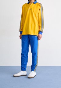 Polo amarillo de manga larga con cuello azul, combinado con pantalones deportivos azules que tienen rayas amarillas. Zapatillas blancas con suelas marrón claro.