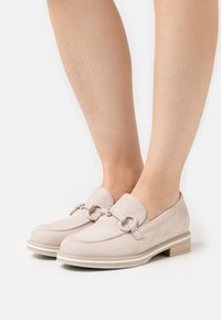 Mocasines de gamuza beige claro con un pequeño tacón cuadrado, que presentan un herraje decorativo de cadena y un pespunte en contraste a lo largo de la suela.