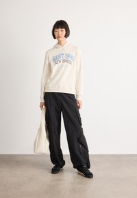Young woman stands wearing a cream hoodie with "GANT 1949" text, black cargo pants, black sneakers, and holding a large cream tote bag.