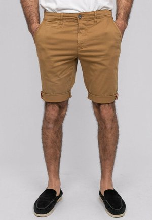 Homme portant un short chino beige retroussé avec poches avant, assorti à des chaussures noires à enfiler, debout devant un fond uni.