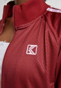 Červená bunda na zip s mřížkovou texturou, žebrovaným límcem a vyšitým logem. Obsahuje bílé pruhy podél ramen.