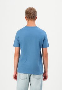 Fiatal férfi, rövid göndör hajjal, kék, rövid ujjú pólót és világos denim farmert viselve, háttal a kamerának áll.