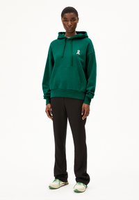Haut à capuche vert avec une poche frontale, arborant un petit logo en ruban blanc. Associé à un pantalon noir et des baskets au style rétro avec des accents verts.