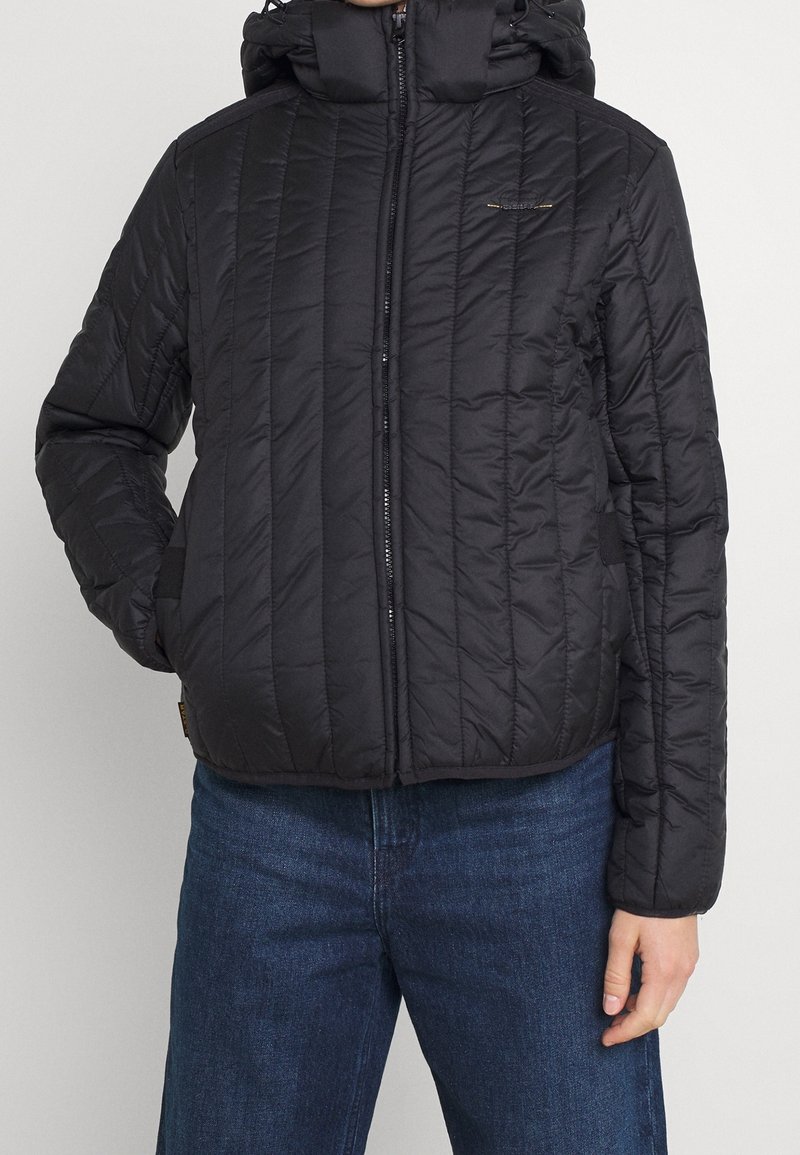 Veste noire matelassée à capuche avec fermeture éclair à l'avant, portée sur un jean bleu, main gauche de la personne dans la poche, main droite détendue sur le côté.