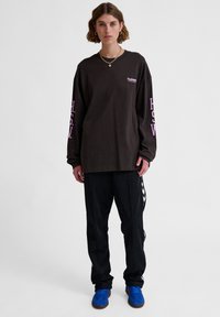 Schwarzes Langarmshirt mit pinkem Logo auf der Brust und an den Ärmeln, kombiniert mit schwarzen Jogginghosen mit einem weißen Chevron-Muster und blauen Schuhen.
