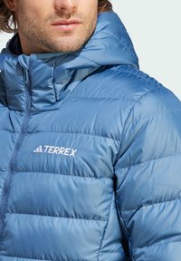 Chaqueta azul acolchada con capucha, que presenta un diseño de acolchado horizontal, cremallera frontal y el logo de Terrex en blanco. Textura suave y brillante.
