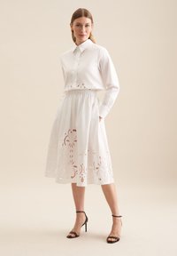 Hvid kjole med kraveskjorte for og en lagdelt nederdel med broderede blomster mønstre. Inkluderer elastisk talje og lommer.