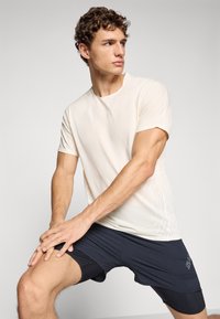 Young man stretching legs wearing a beige athletic t-shirt and black layered shorts against a plain light background.