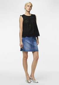 Top en dentelle noire avec une finition en scallop, associé à une jupe en denim. La mannequin porte des talons argentés. Silhouette simple et décontractée avec des détails au niveau du décolleté.