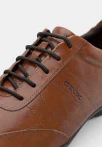 Brown leather shoe featuring black laces, textured surface, and stitched accents. "GEOX" logo embossed on the side.