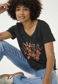 T-shirt noir à col en V avec un motif floral dans des nuances d'orange et de marron. Dispose de manches courtes et d'une coupe décontractée, associé à un jean bleu clair.