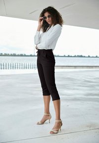 White long-sleeve blouse, black tailored cropped pants, and beige high-heeled sandals. Large round sunglasses. Smooth textures, fitted design.