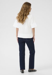 Top bianco a coste con maniche corte e larghe, abbinato a jeans blu scuro e scarpe nere decorate con borchie argentate, visto da dietro.