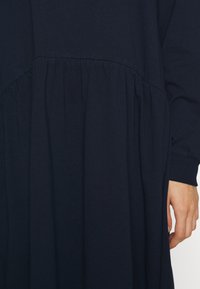 Námořnické modré šaty mají řasený panel v pase, měkkou bavlněnou látku a dlouhé rukávy se zúženým manžetou.