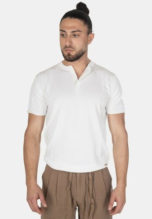 Uomo con capelli scuri raccolti in uno chignon che indossa una maglia henley bianca a maniche corte e pantaloni plissettati marrone chiaro, guarda leggermente verso il basso a sinistra.