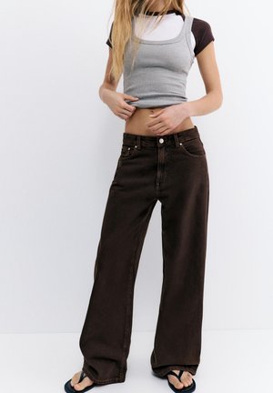 Femme portant des hauts superposés gris et noirs avec un jean marron foncé taille haute et jambes larges, ainsi que des tongs noires, debout devant un fond blanc.