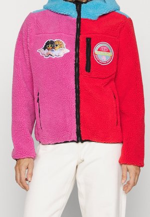 Chaqueta de forro polar con el lado izquierdo rosa, el lado derecho rojo, cuello azul, dos parches: un par de ángeles con gafas de sol y un parche redondo con logo.