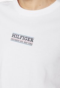Valkoinen crewneck-paita, jossa on musta teksti "HILFIGER COLUMBUS AVE. NEW YORK" sinisen ja punaisen alleviivauksen kanssa rinnassa.