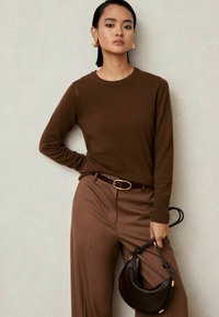 Suéter marrón de punto con escote redondo, combinado con pantalones marrones de cintura alta, un bolso oscuro y grandes aretes de aro dorados.